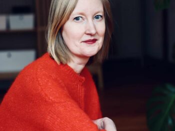 Eine Frau mit schulterlangem Haar, die einen knallroten Pullover trägt, blickt in die Kamera.