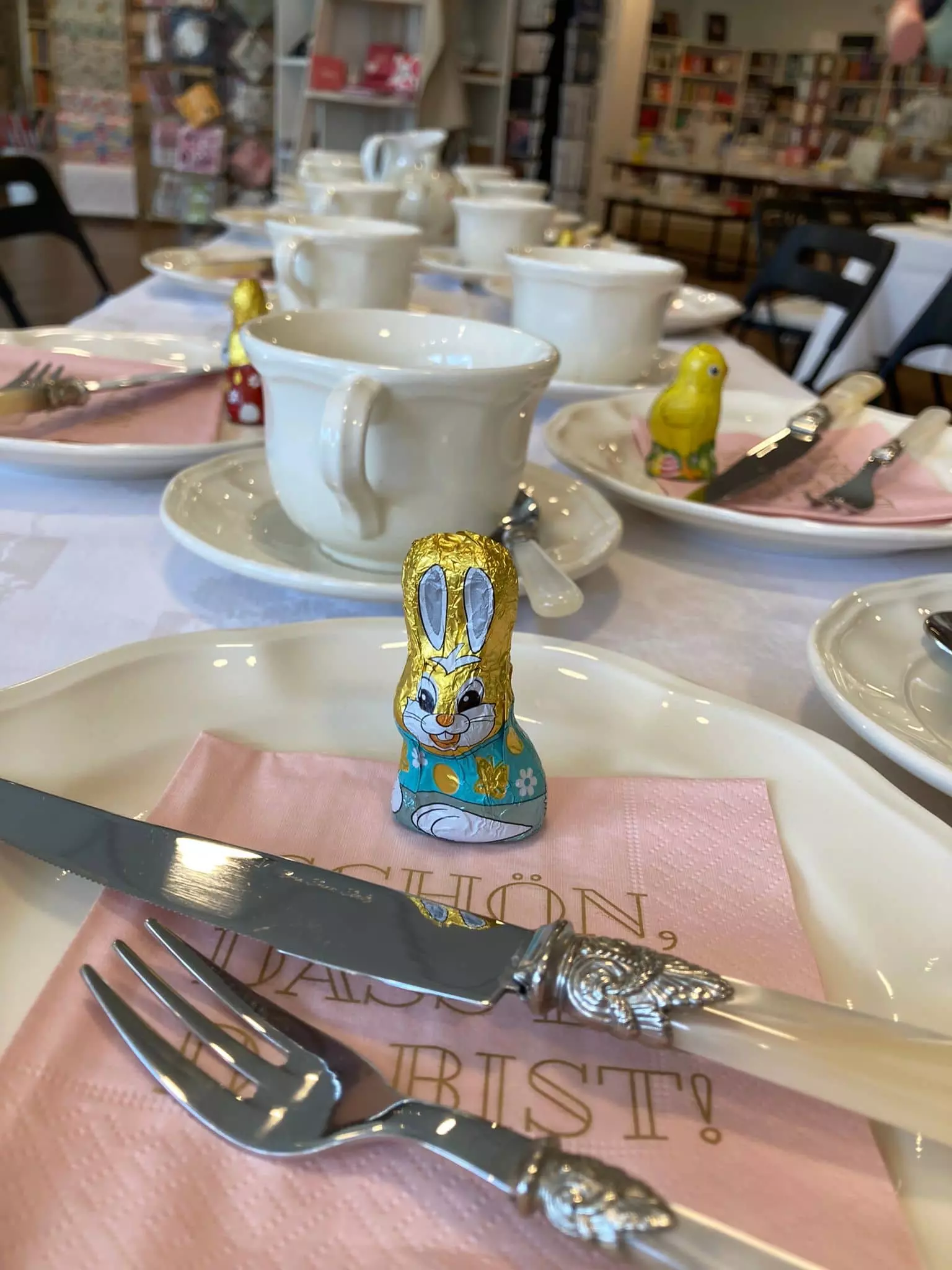 Ein Tischgedeck für eine Teeparty mit weißen Tassen und Tellern, silbernem Besteck und rosa Servietten. Auf jedem Teller liegen eingewickelte Schokoladenhasen und -küken. Im Hintergrund sind Regale mit Büchern zu sehen.