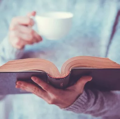 Eine Frau hält ein aufgeschlagenes Buch und eine Tasse Kaffee.
