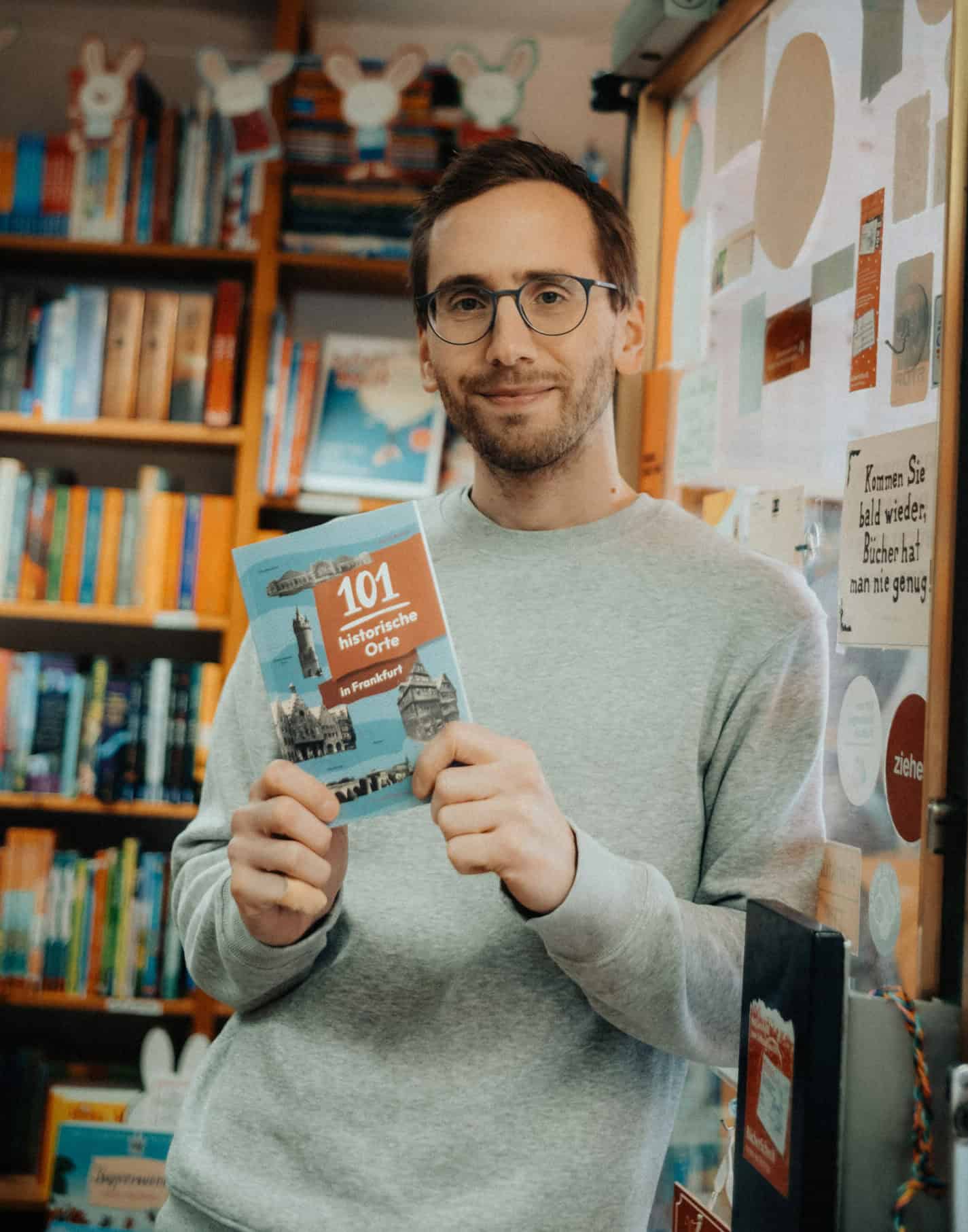 Ein Mann mit Brille und grauem Pullover steht in einer Buchhandlung und hält ein Buch mit dem Titel „101 Dinge, die man in der Schweiz tun kann“ in der Hand.