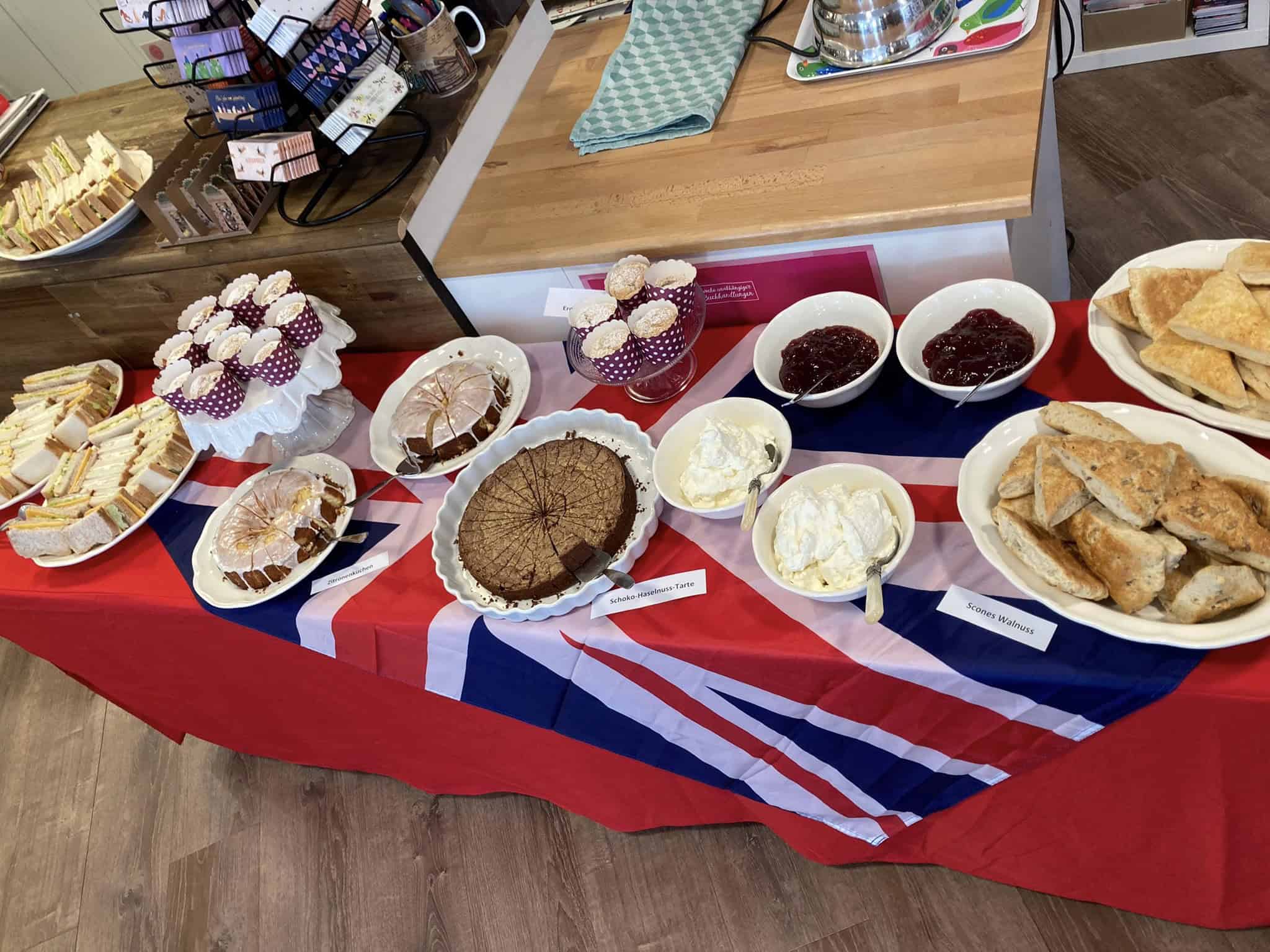 Ein mit einer Tischdecke im Union-Jack-Stil bedeckter Tisch, auf dem verschiedene Lebensmittel, darunter Sandwiches, Scones, Kuchen und Gewürze, präsentiert werden.