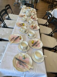 Ein langer Tisch ist mit weißen Teetassen, Tellern und Servietten zum Tee gedeckt. Auf jedem Gedeck steht eine kleine gelbe und rote Puppe. Weiße Tischdecken und schwarze Klappstühle runden das Arrangement ab.