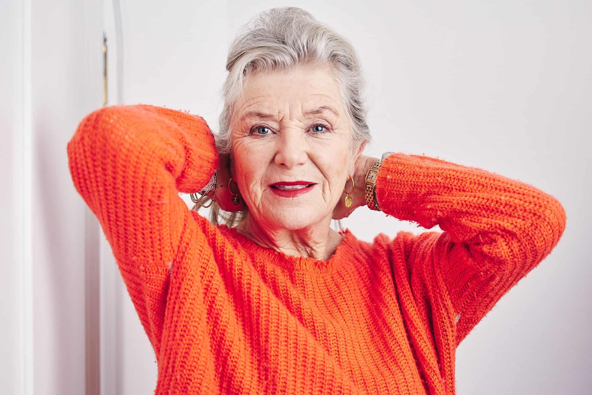 Eine ältere Frau mit grauem Haar und rotem Lippenstift sowie einem orangefarbenen Strickpullover posiert mit den Händen hinter dem Kopf vor einem weißen Hintergrund.