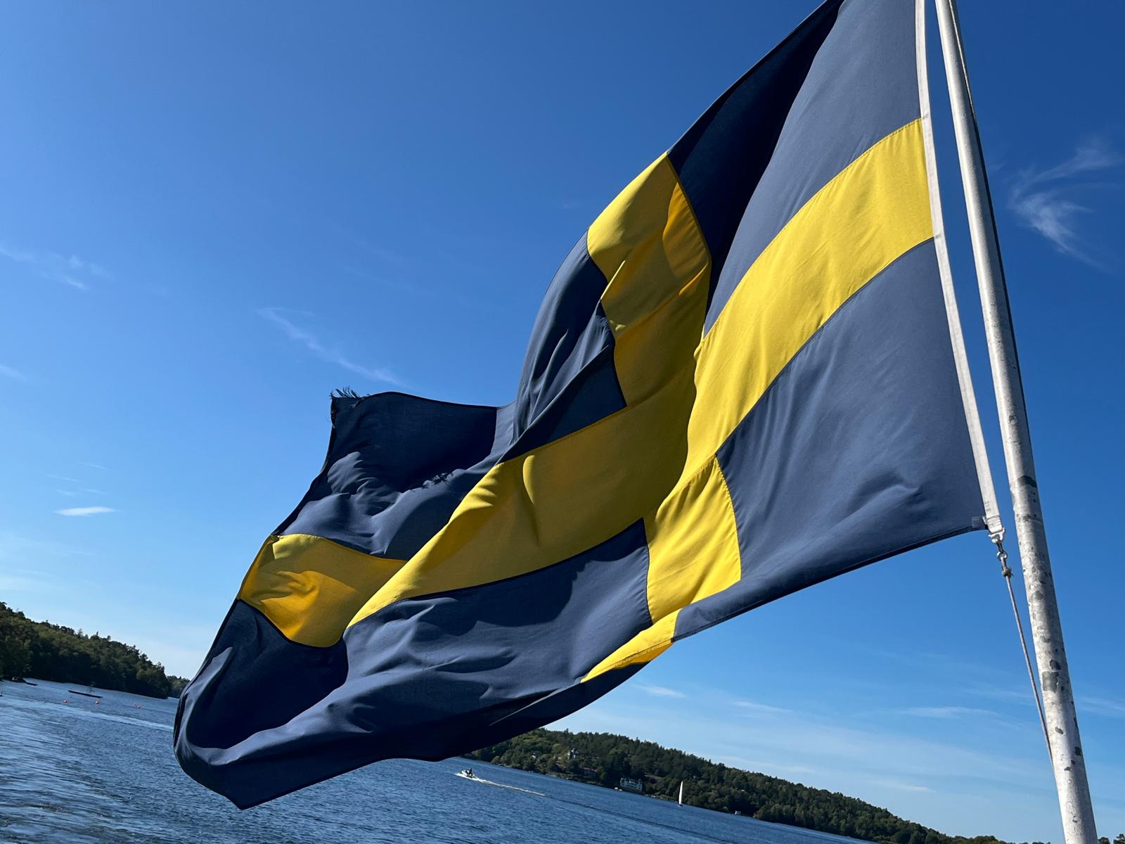 Schwedische Flagge weht an einer Stange vor dem Hintergrund eines klaren blauen Himmels und Wassers.