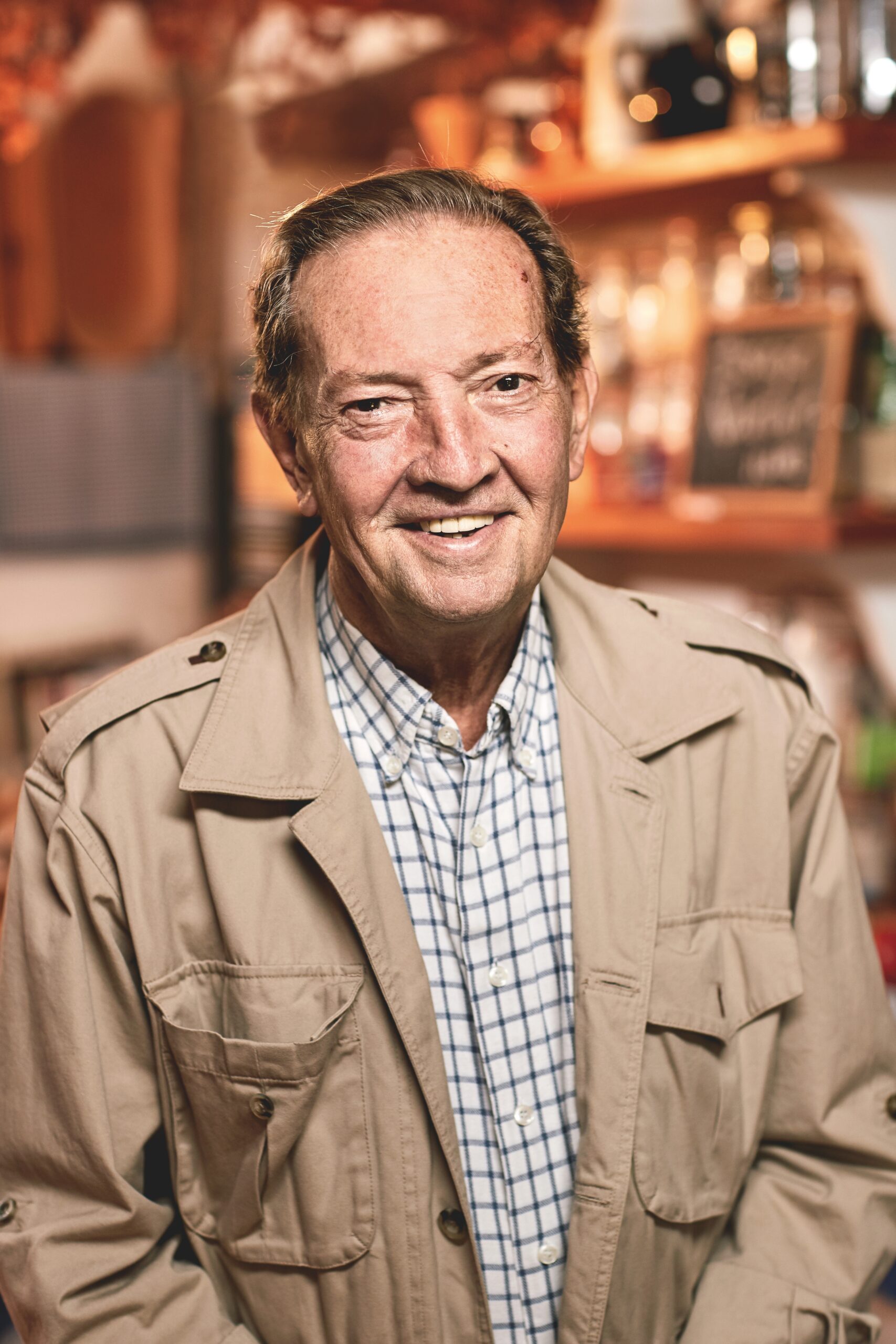 Ein Mann in beiger Jacke und kariertem Hemd steht lächelnd im Haus. Im unscharfen Hintergrund sind Regale mit Flaschen und eine Tafel zu sehen.