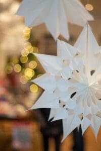 Im Vordergrund hängen weiße Papiersterne, im Hintergrund sind verschwommene gelbe Weihnachtslichter und undeutliche Innenobjekte zu sehen.