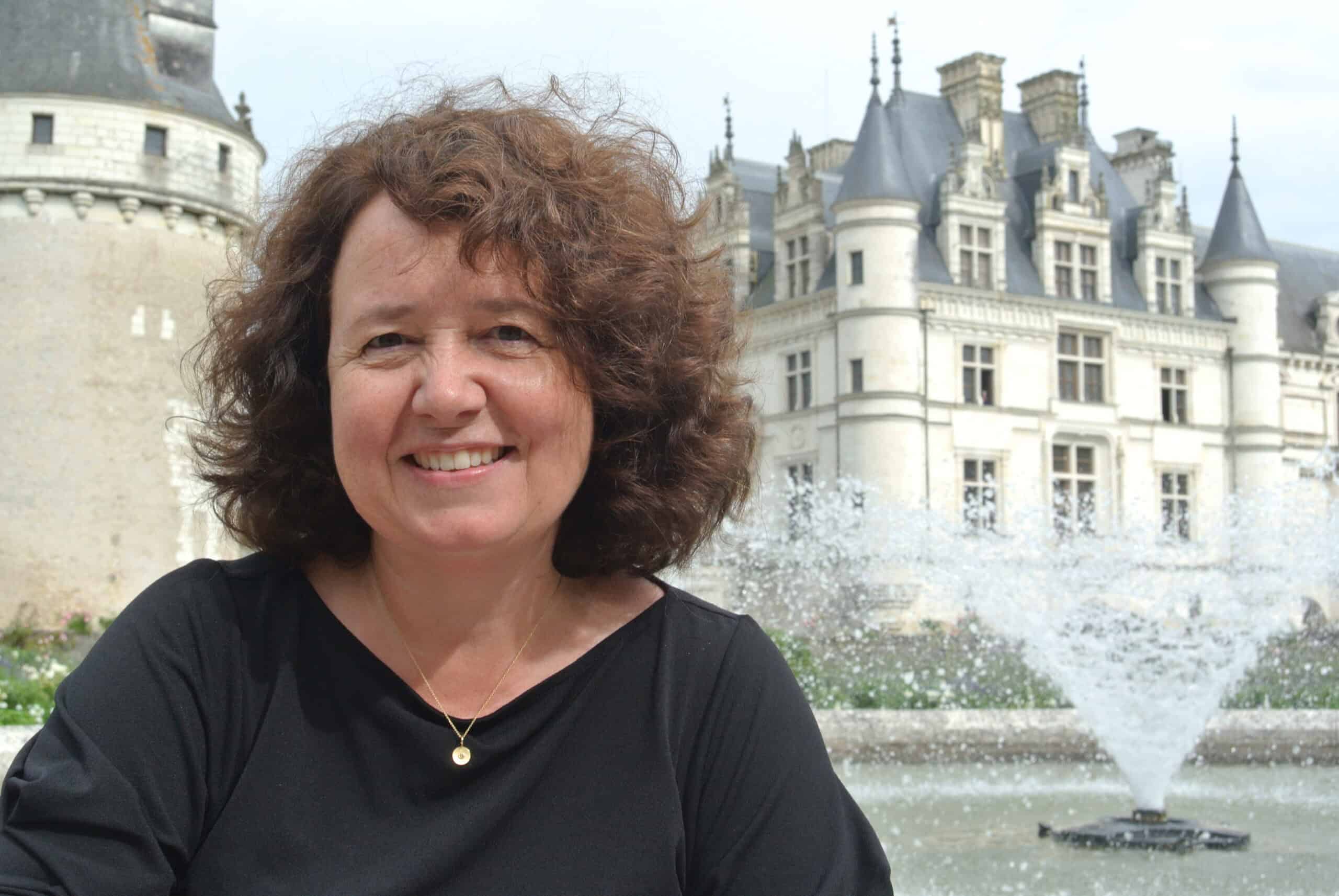 Eine Frau mit lockigem braunem Haar lächelt vor einem historischen Gebäude im europäischen Stil und einem spritzenden Brunnen.