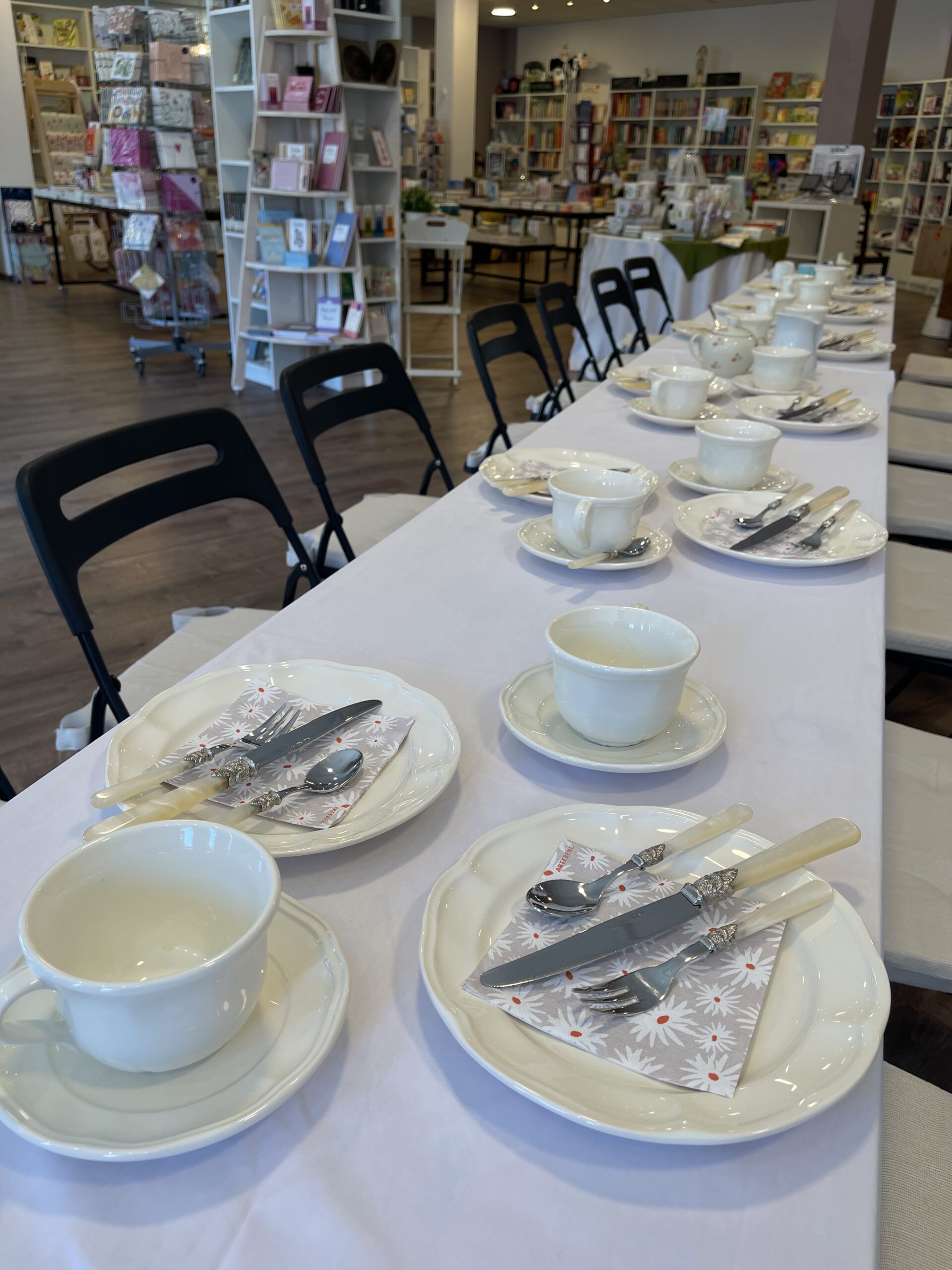 Ein langer, mit weißen Teetassen, Untertassen, Tellern und Besteck auf Servietten gedeckter Tisch, arrangiert in einem Laden, im Hintergrund sind Bücher und Geschenke ausgestellt.