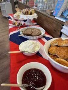 Auf einem Tisch mit einer Tischdecke mit der britischen Flagge stehen Scones, Marmelade, Streichrahm, Kuchen und Gebäck, im Hintergrund sind Besteck und Bücher zu sehen.