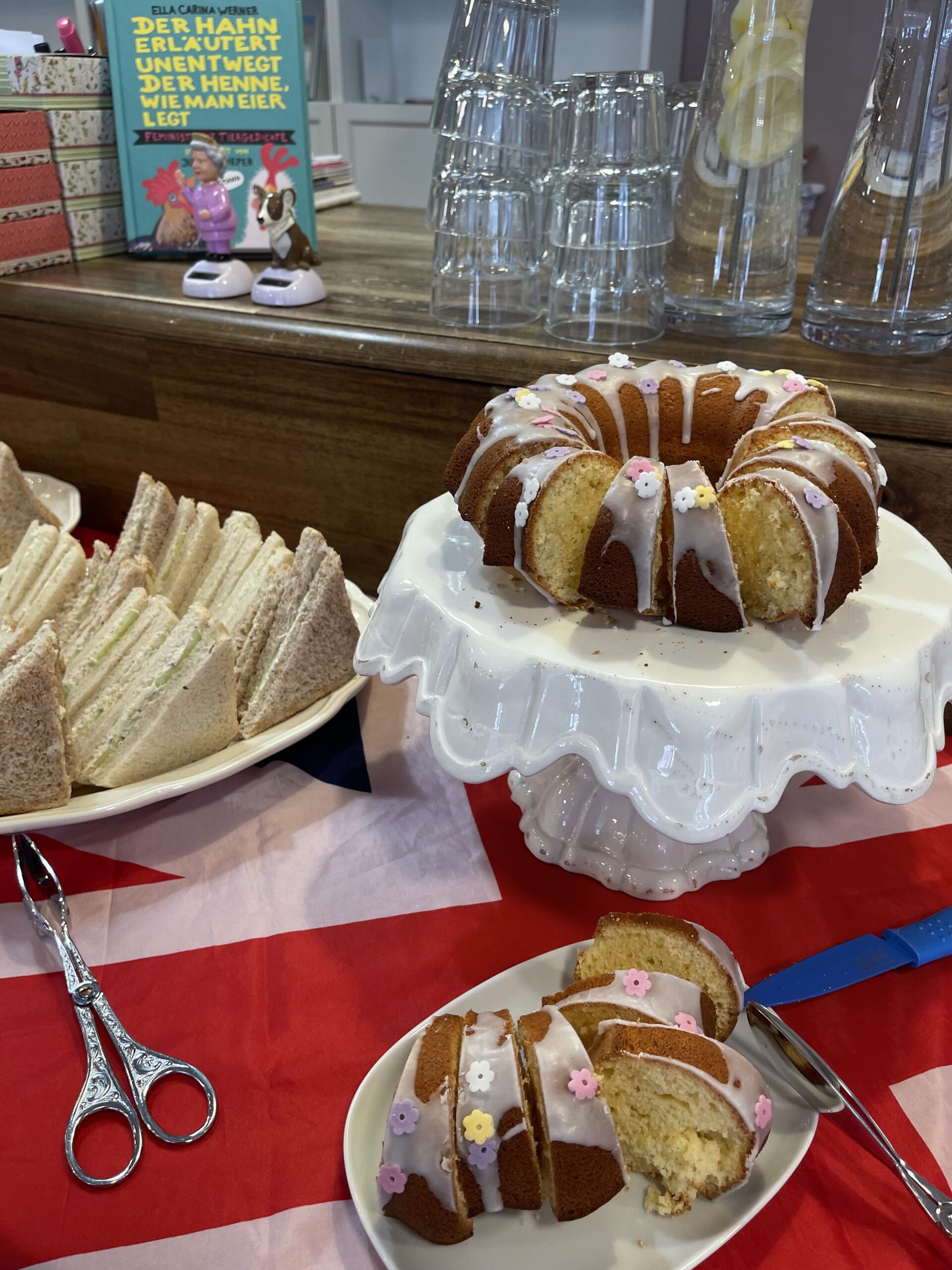 Ein angeschnittener Gugelhupf mit rosa und weißem Zuckerguss steht auf einem Tortenständer neben einem Teller mit verschiedenen Sandwichdreiecken auf einem Tisch mit einer rot-weißen Tischdecke.
