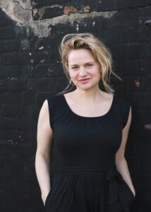 Eine Frau mit blondem Haar und einem schwarzen ärmellosen Kleid steht vor einer dunklen, strukturierten Wand und blickt mit neutralem Gesichtsausdruck in die Kamera.