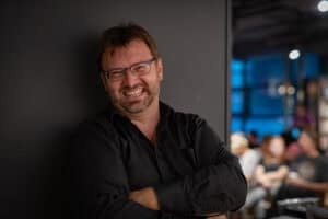 Ein Mann mit Brille, Bart und schwarzem Hemd steht mit verschränkten Armen im Haus und lächelt in die Kamera. Menschen und Lichter im Hintergrund sind unscharf.