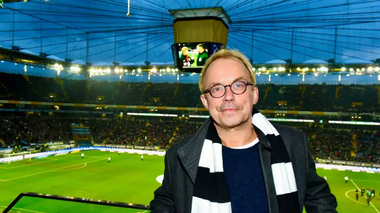 Ein Mann mit Brille und gestreiftem Schal steht während eines Abendspiels in einem großen Fußballstadion voller Zuschauer.