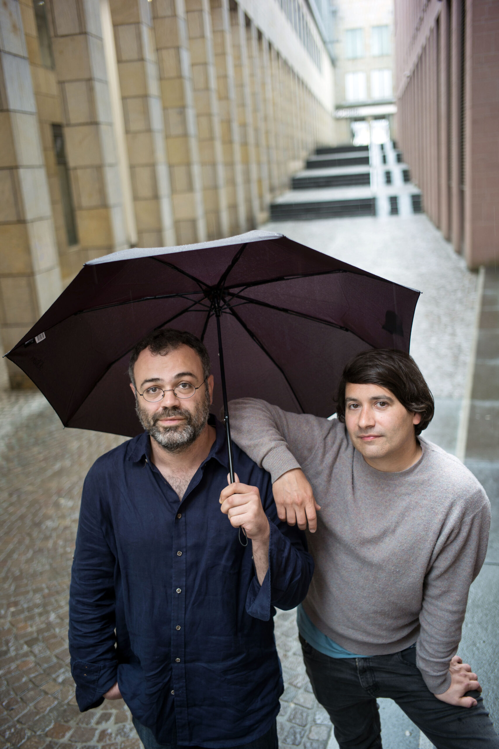 Zwei Männer stehen zusammen auf einem schmalen Gehweg im Freien; einer hält einen schwarzen Regenschirm, während sich der andere auf seine Schulter stützt. Der Boden ist nass, was auf kürzlichen Regen hindeutet.