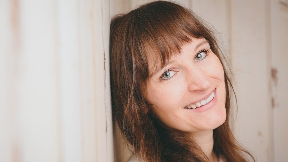 Eine Frau mit hellen Augen und glattem braunem Haar mit Pony lehnt lächelnd ihren Kopf an eine helle Holzwand.