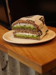 Ein Sandwich mit dunklem Brot, Frischkäse und Gurkenscheiben wird halbiert und auf einen weißen Teller gelegt.