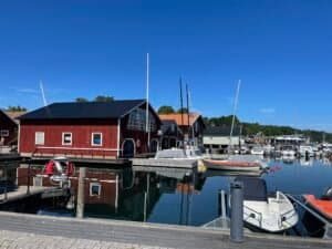 Kleine Boote, die in einem Jachthafen angedockt sind, mit roten Holzboothäusern, die sich im ruhigen Wasser unter einem klaren blauen Himmel spiegeln.
