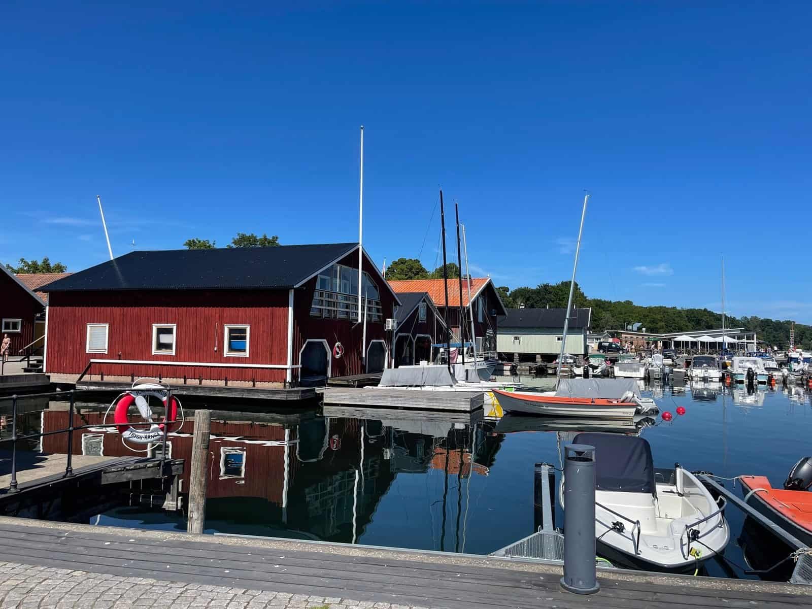 Kleine Boote, die in einem Jachthafen angedockt sind, mit roten Holzboothäusern, die sich im ruhigen Wasser unter einem klaren blauen Himmel spiegeln.