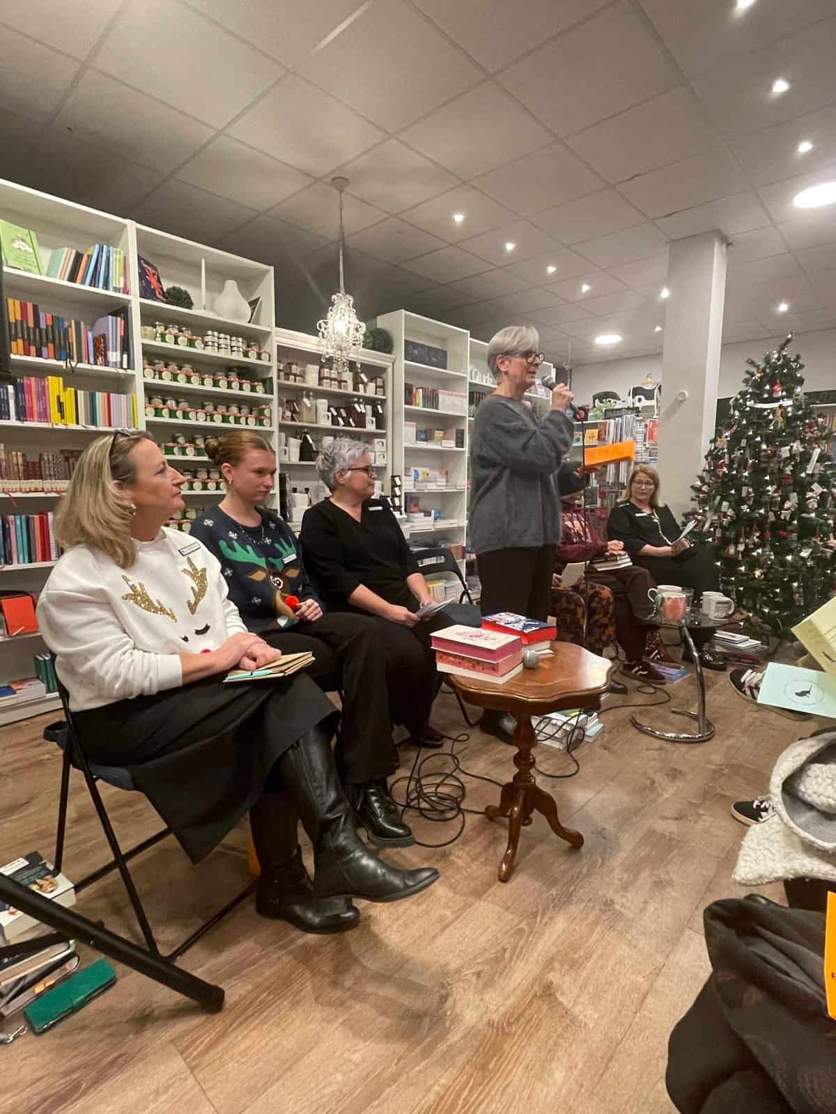 Vier Frauen sitzen in einer Buchhandlung, eine von ihnen steht und spricht in ein Mikrofon. Im Hintergrund sind Bücher und ein geschmückter Weihnachtsbaum zu sehen.