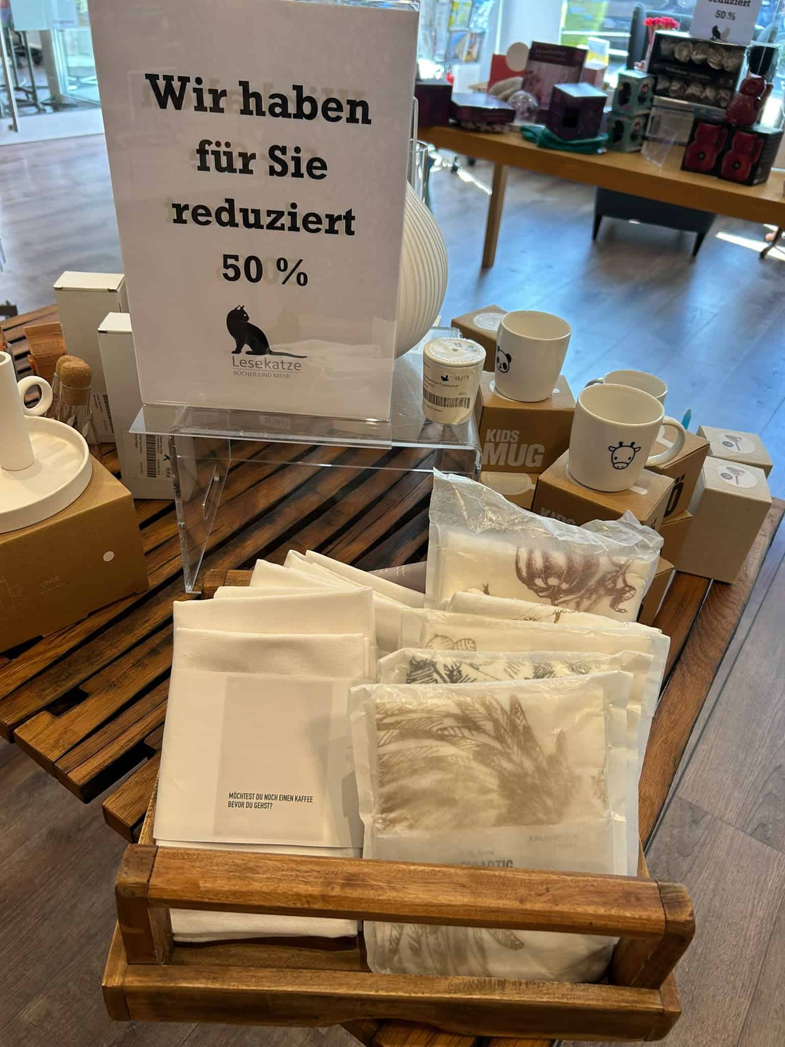Eine Verkaufsauslage mit Servietten und Tüchern in einer Holzschale. Auf einem Schild in deutscher Sprache steht: „Wir haben für Sie 50 % reduziert“, was auf einen Rabatt von 50 % hinweist. Im Hintergrund sind weitere Geschenkartikel zu sehen.