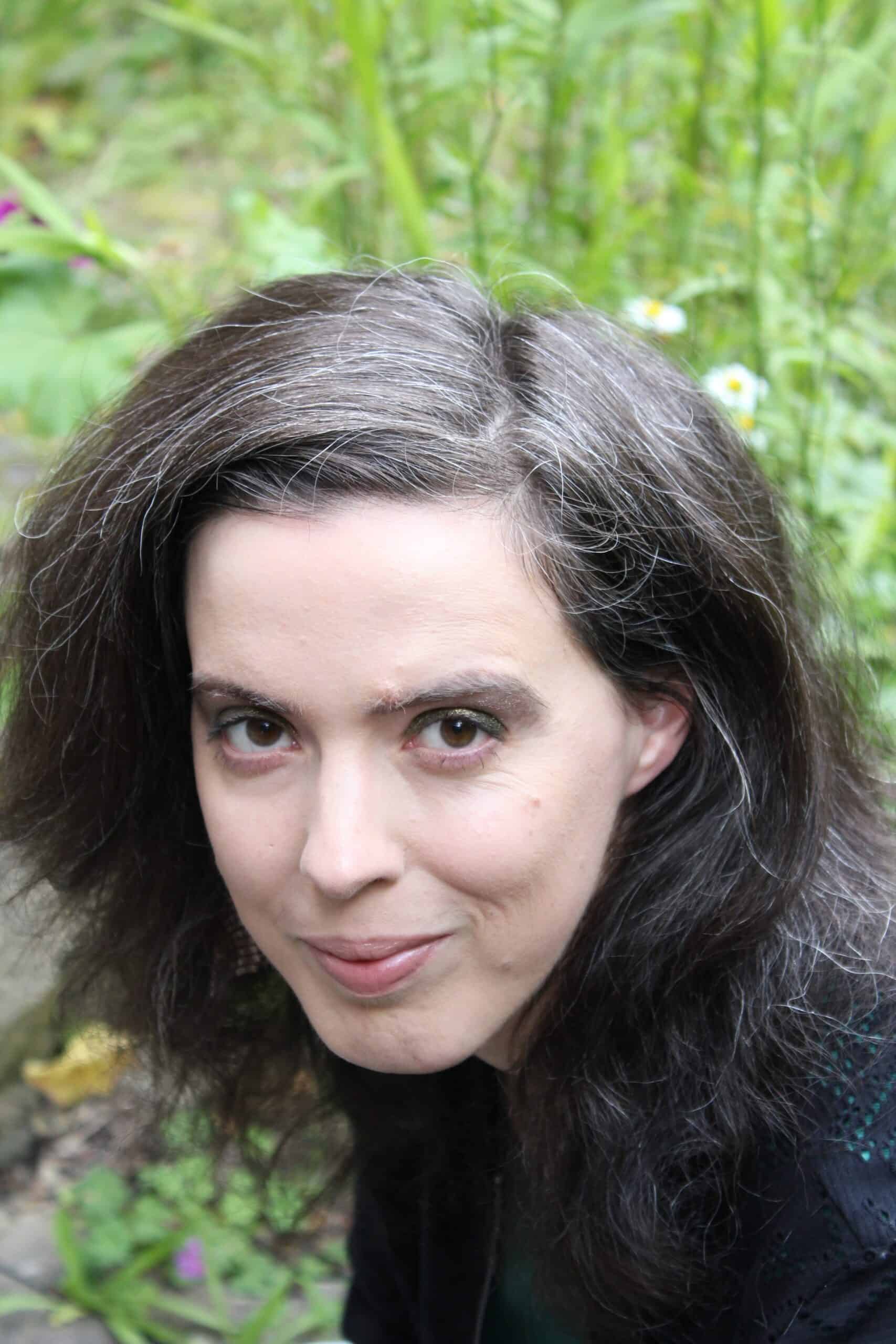 Eine Person mit langen, dunklen Haaren und einigen grauen Strähnen blickt in die Kamera; im Hintergrund sind im Freien Grünpflanzen und Blumen zu sehen.