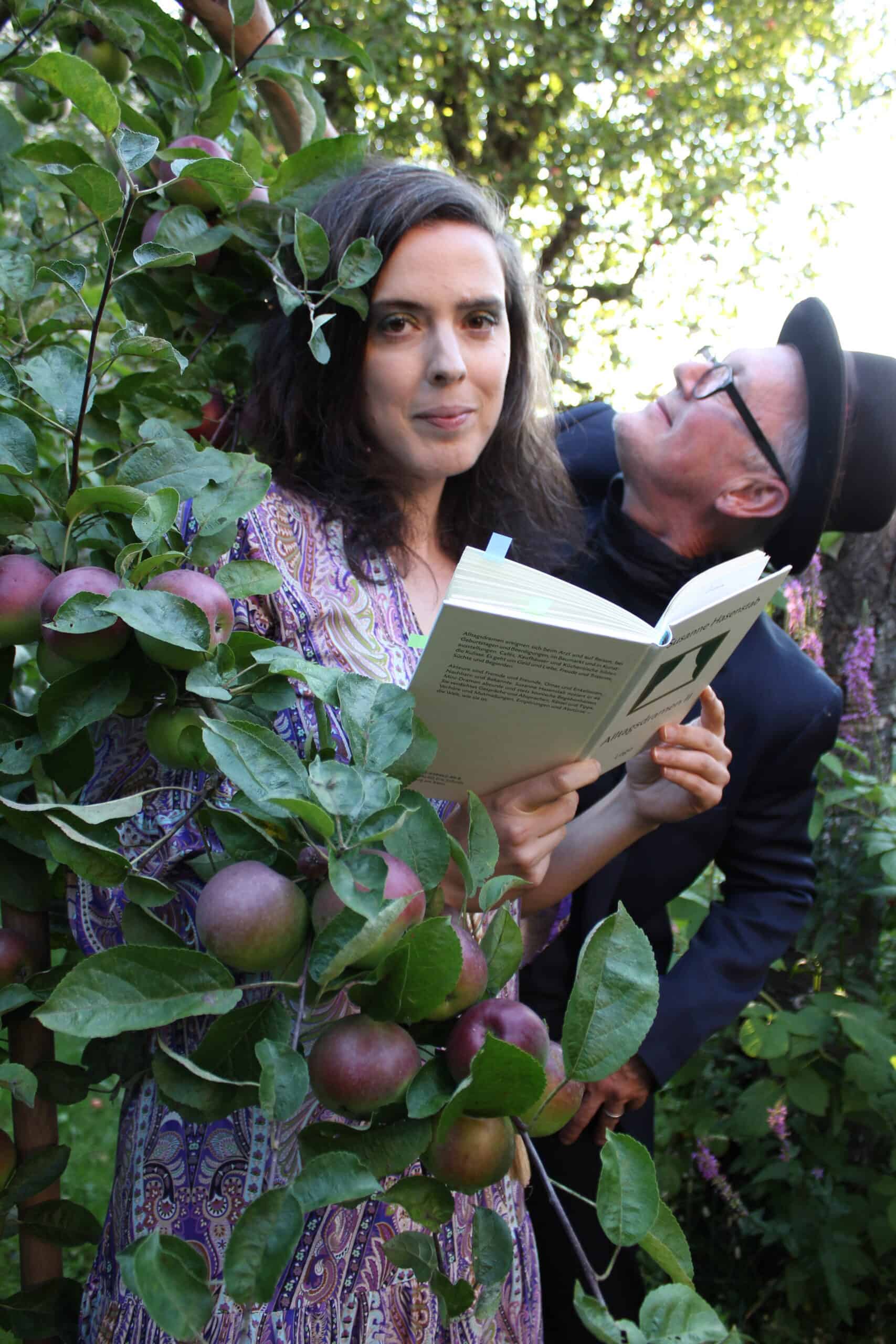 Eine Frau steht zwischen den Ästen eines Apfelbaums und liest ein Buch, während ein Mann mit Hut und Brille sie von hinten ansieht.