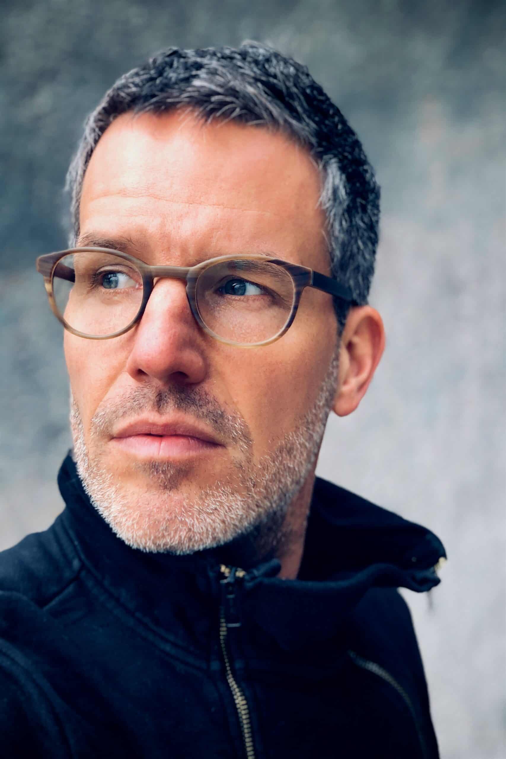 Ein Mann mit kurzen grauen Haaren, Bartstoppeln und Brille, der eine dunkle Jacke mit Reißverschluss trägt, blickt vor einem verschwommenen grauen Hintergrund zur Seite.