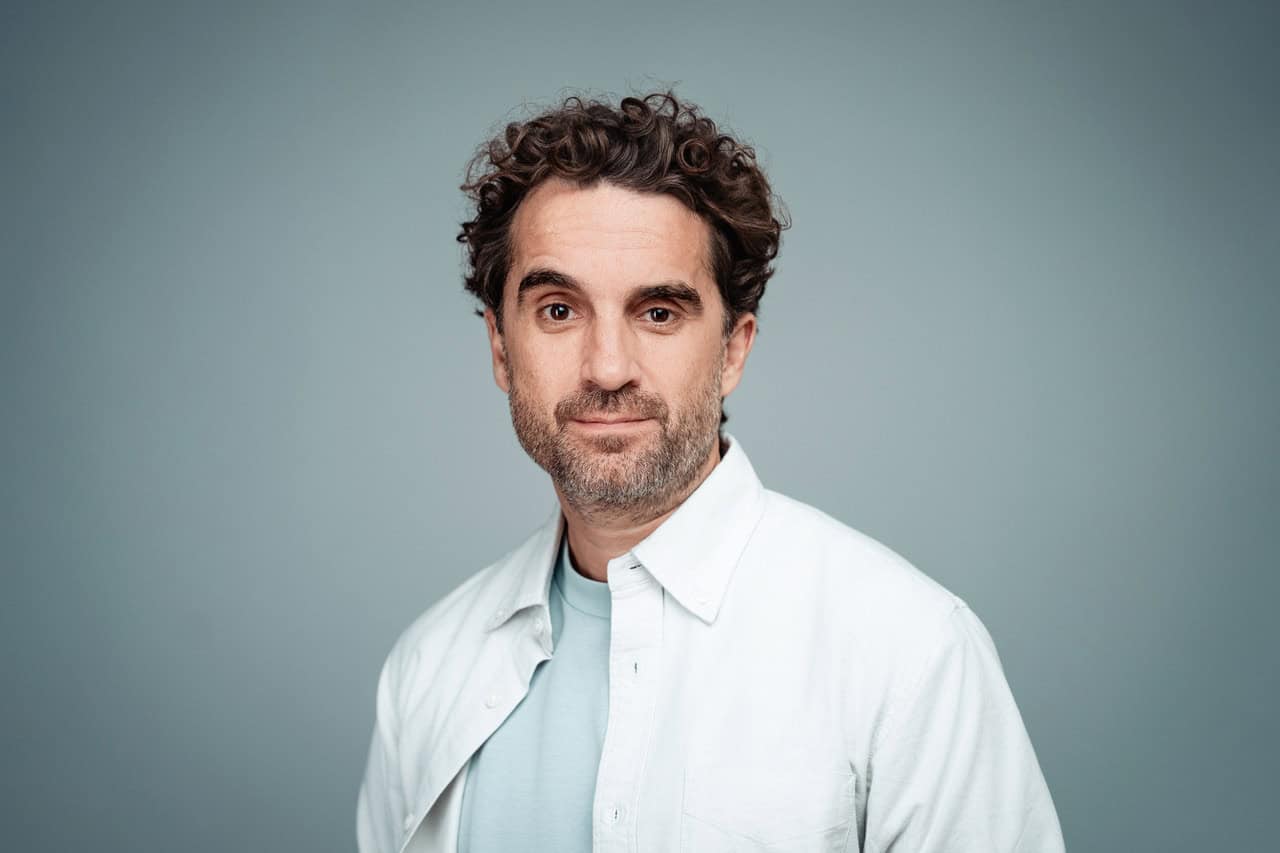 Ein Mann mit lockigem braunem Haar und Bart, der ein weißes Hemd über einem hellblauen T-Shirt trägt und vor einem schlichten grauen Hintergrund steht.