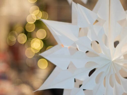 Im Vordergrund hängen weiße Papiersterne, im Hintergrund sind verschwommene gelbe Weihnachtslichter und undeutliche Innenobjekte zu sehen. {{brizy_dc_image_alt entityId=