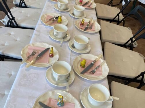 Ein langer Tisch ist mit weißen Teetassen, Tellern und Servietten zum Tee gedeckt. Auf jedem Gedeck steht eine kleine gelbe und rote Puppe. Weiße Tischdecken und schwarze Klappstühle runden das Arrangement ab. {{brizy_dc_image_alt entityId=