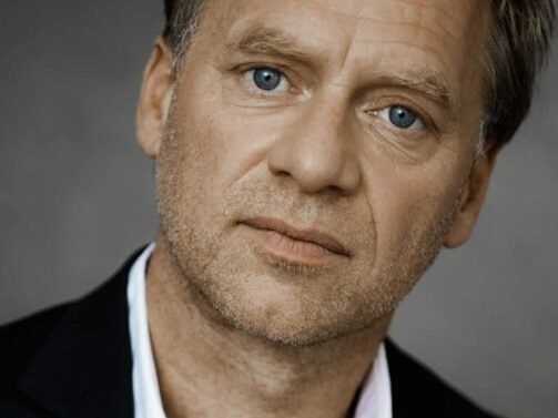 Ein Mann mit hellem Haar, blauen Augen und hellem Bart, der einen schwarzen Blazer über einem weißen Hemd trägt, blickt vor einem neutralen Hintergrund direkt in die Kamera.