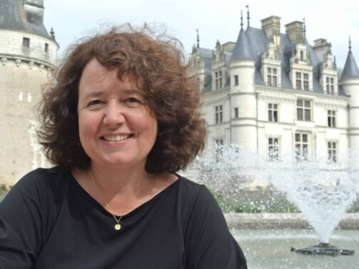 Eine Frau mit lockigem braunem Haar lächelt vor einem historischen Gebäude im europäischen Stil und einem spritzenden Brunnen.