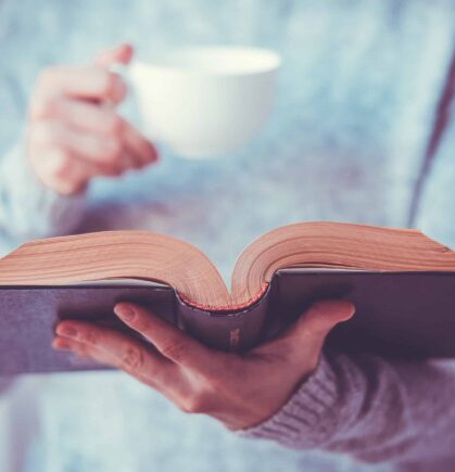 Eine Frau hält ein aufgeschlagenes Buch und eine Tasse Kaffee.