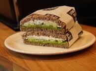 Ein Sandwich mit dunklem Brot, Frischkäse und Gurkenscheiben wird halbiert und auf einen weißen Teller gelegt.