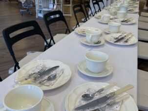 IMG_5439 Ein langer, mit weißen Teetassen, Untertassen, Tellern und Besteck auf Servietten gedeckter Tisch, arrangiert in einem Laden, im Hintergrund sind Bücher und Geschenke ausgestellt.