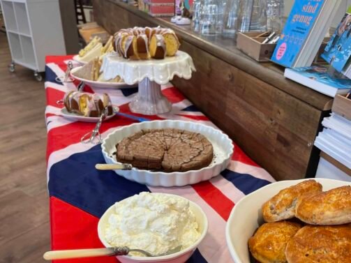 Auf einem Tisch mit einer Tischdecke mit der britischen Flagge stehen Scones, Marmelade, Streichrahm, Kuchen und Gebäck, im Hintergrund sind Besteck und Bücher zu sehen. {{brizy_dc_image_alt entityId=