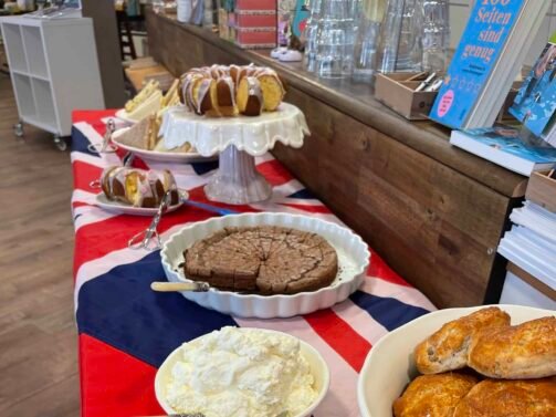 Auf einem Tisch mit einer Tischdecke mit der britischen Flagge stehen Scones, Marmelade, Streichrahm, Kuchen und Gebäck, im Hintergrund sind Besteck und Bücher zu sehen. {{brizy_dc_image_alt entityId=