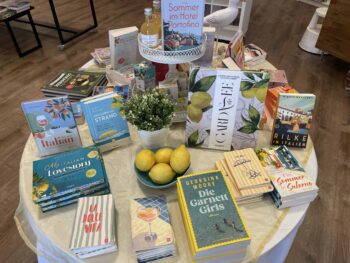 Auf einem runden Tisch sind Bücher mit sommerlichen Themen, eine Topfpflanze, Zitronen, Wein und Zeitschriften ausgestellt, die in einer Buchhandlung auf einem Holzboden aufgestellt sind.