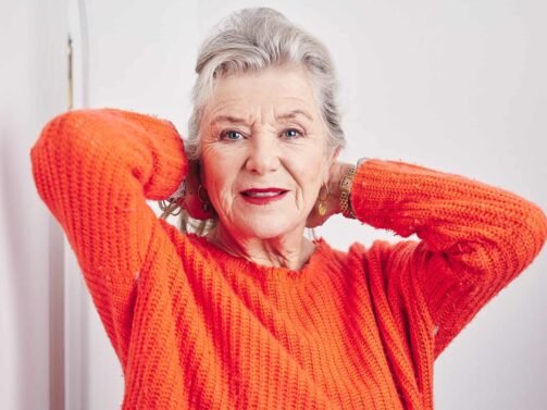 Eine ältere Frau mit grauem Haar und rotem Lippenstift sowie einem orangefarbenen Strickpullover posiert mit den Händen hinter dem Kopf vor einem weißen Hintergrund.