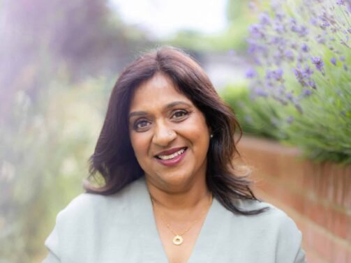 Eine Frau mit dunklem Haar, einer hellgrünen Bluse und einem gemusterten Rock hält eine Messingschale, während sie im Freien neben einer Backsteinmauer und Lavendelpflanzen sitzt.