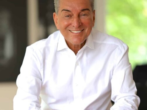 Ein Mann in einem weißen Hemd lächelt, während er sich auf die Lehne eines roten Stuhls stützt. Der Hintergrund ist unscharf, und durch ein Fenster ist grünes Laub zu sehen. {{brizy_dc_image_alt entityId=