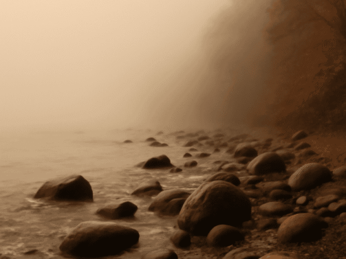 Felsiges Ufer mit großen Steinen neben ruhigem Wasser, eingehüllt in dichten Nebel, mit Herbstbäumen am rechten Rand. {{brizy_dc_image_alt entityId=