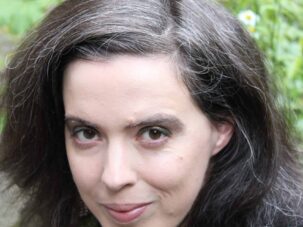 Portraet_Susanne1 Eine Person mit langen, dunklen Haaren und einigen grauen Strähnen blickt in die Kamera; im Hintergrund sind im Freien Grünpflanzen und Blumen zu sehen.