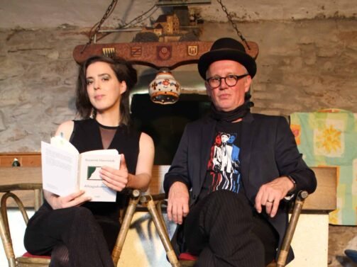 Presse1_SusanneEmil Zwei Personen sitzen auf hohen Stühlen, eine hält ein offenes Buch und die andere trägt Hut und Brille, in einem rustikalen Innenraum.