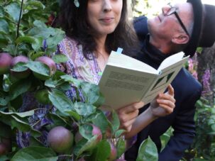 Presse3_SusanneEmil Eine Frau steht zwischen den Ästen eines Apfelbaums und liest ein Buch, während ein Mann mit Hut und Brille sie von hinten ansieht.