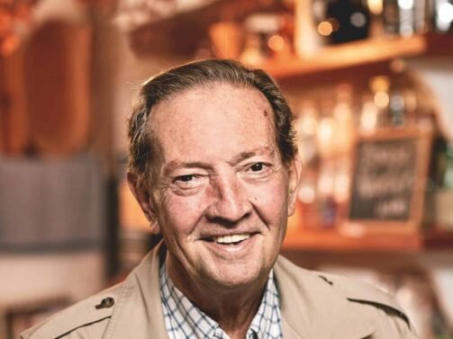 Ein Mann in beiger Jacke und kariertem Hemd steht lächelnd im Haus. Im unscharfen Hintergrund sind Regale mit Flaschen und eine Tafel zu sehen.