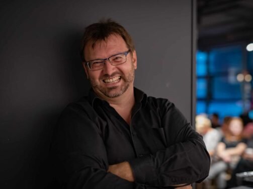 Ein Mann mit Brille, Bart und schwarzem Hemd steht mit verschränkten Armen im Haus und lächelt in die Kamera. Menschen und Lichter im Hintergrund sind unscharf.