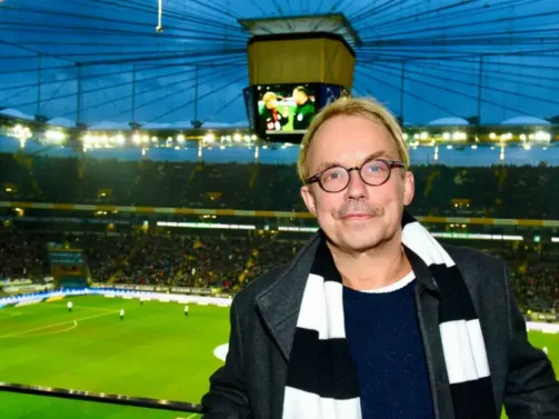 d7e90de67d2354dff007e6221c1ebad7850e5b Ein Mann mit Brille und gestreiftem Schal steht während eines Abendspiels in einem großen Fußballstadion voller Zuschauer.