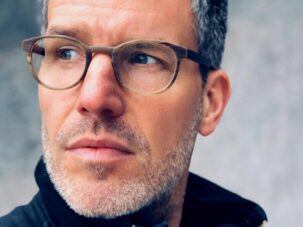 Image0 Ein Mann mit kurzen grauen Haaren, Bartstoppeln und Brille, der eine dunkle Jacke mit Reißverschluss trägt, blickt vor einem verschwommenen grauen Hintergrund zur Seite.
