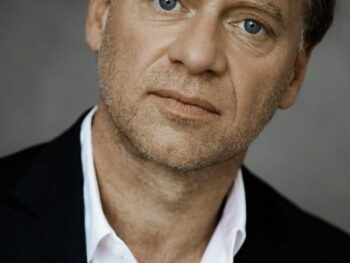 Nahaufnahme eines Mannes mittleren Alters mit hellen Augen, der ein weißes Hemd und einen schwarzen Blazer trägt.